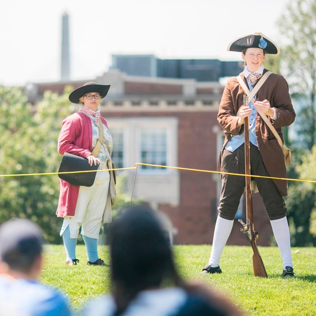 two rangers dressed in colonial era kit speaking to kids sitting on the ground 