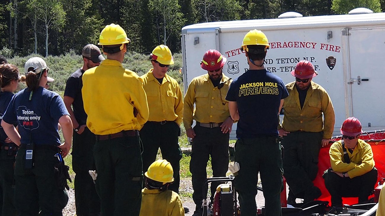 Several people in blue and yellow shirts listen to a man talking to them in front of a white trailer