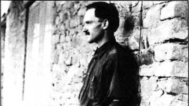 A black and white photo of a man learning against a ruin wall.