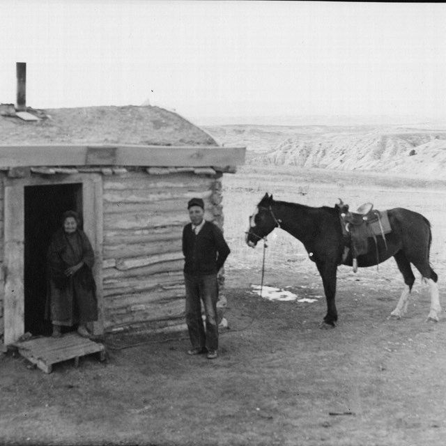 a man, woman, and horse stand in front of a simple house with badlands in the background