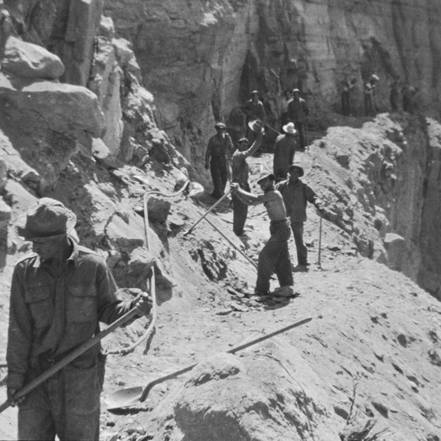 A black and white image of men holding tools working on a rocky trail.