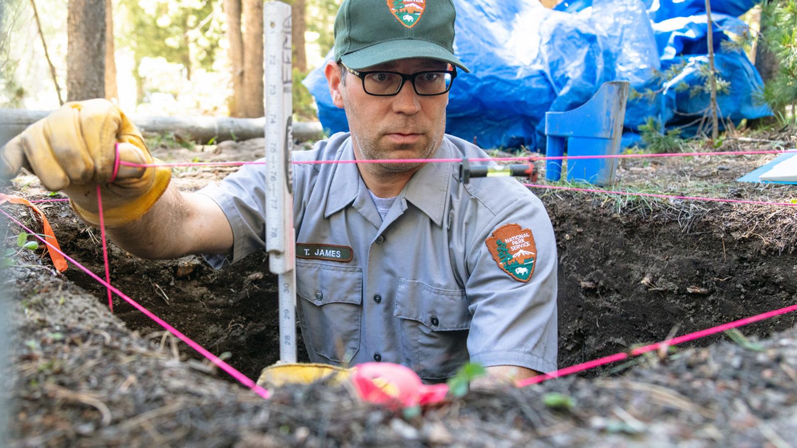 Man in NPS uniform stands in pit, measuring depth with string and ruler