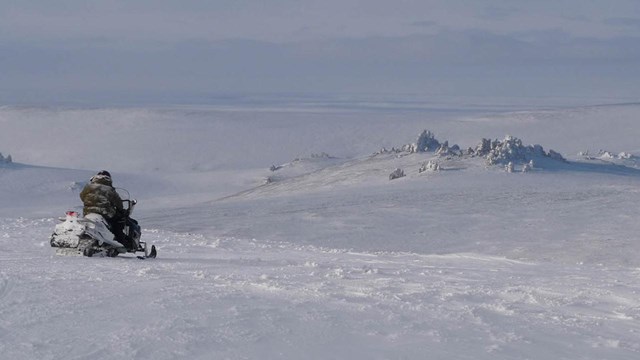 A snowmachiner driving towards snow covered tors in the distance. 