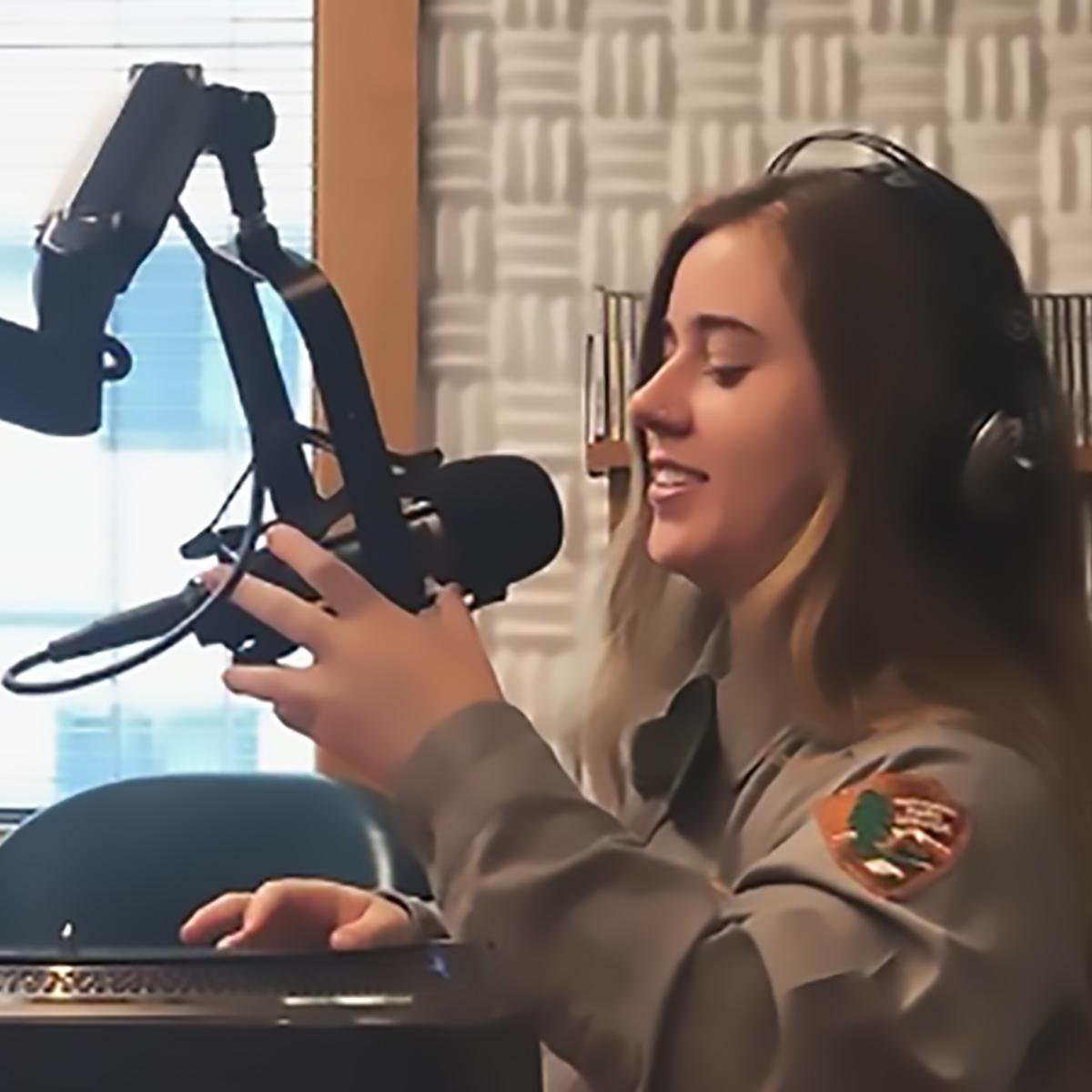 A ranger with headphones on sits at a desk, holding a mirophone.