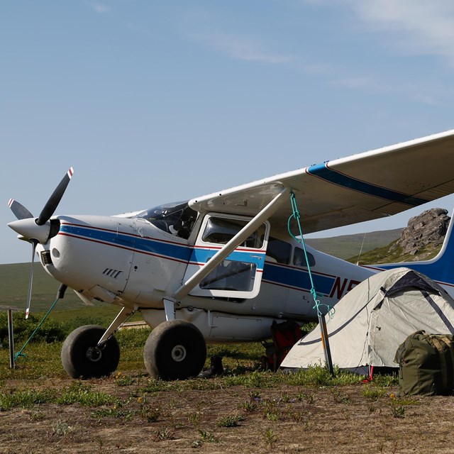 A small airplane parked on the tundra with a gray tent under the wing.