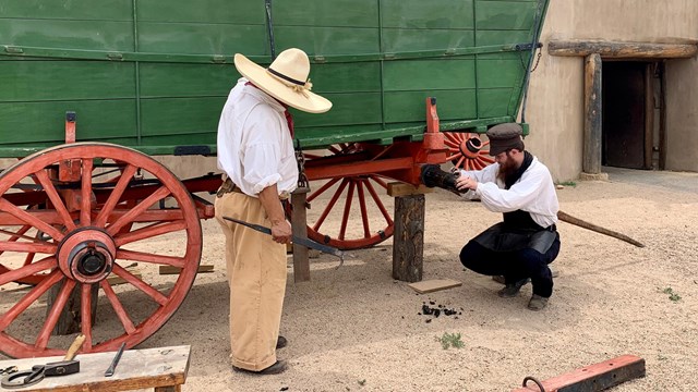 Two men work on a wagon wheel in front of the fort. 