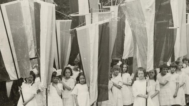 A group of girls in white dresses carrying tri-colored suffrage banners