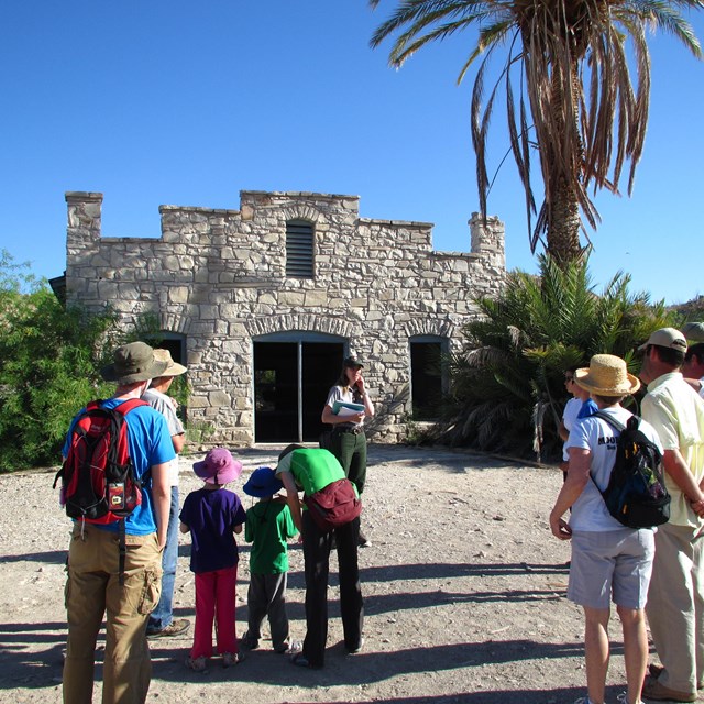 A group of visitors standing in front of a historic structure. A park ranger is pointing at a tree.
