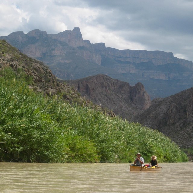 Two people in a canoe floating down the Rio Grande toward mountains covered in foliage.