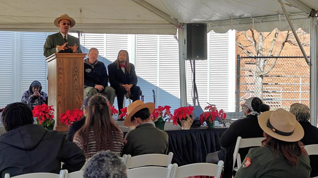 A man stands at a podium on a stage. Three people sit behind him. Rows of people sit facing him.