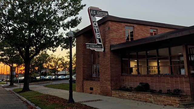 An illuminated neon sign hangs from a two-story brick building. The words "A.G. Gaston Motel" glow.