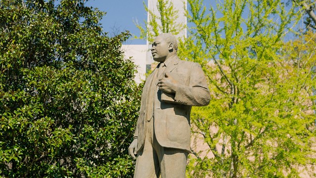 A statue of Dr. King on a pedestal. Trees and a tall building are in the background.