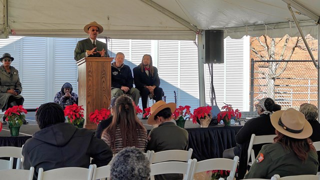 A man stands on stage behind a podium. Three people sit behind him. Rows of people sit facing him.
