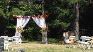 White curtained wedding arch hangs below trees with several seats in field