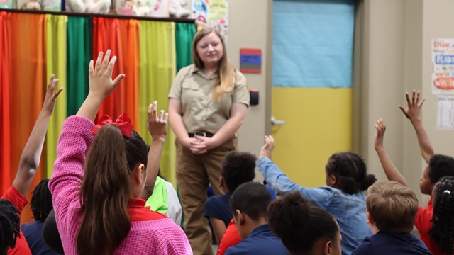 A group of seated students raise their hand to answer a ranger's question during a classroom program