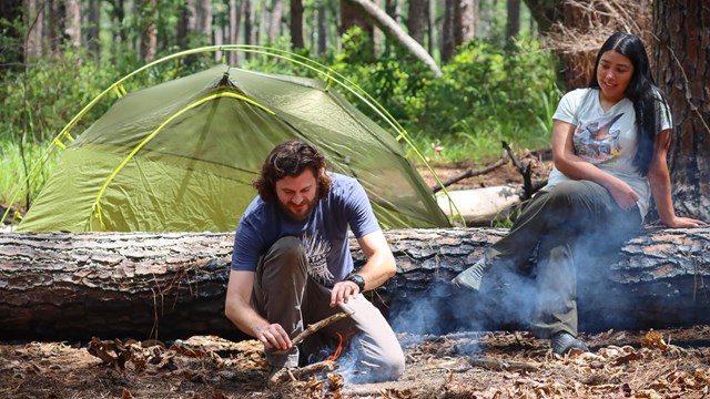 A kneeling man starting a campfire next to a woman sitting on a log in front of a green tent.