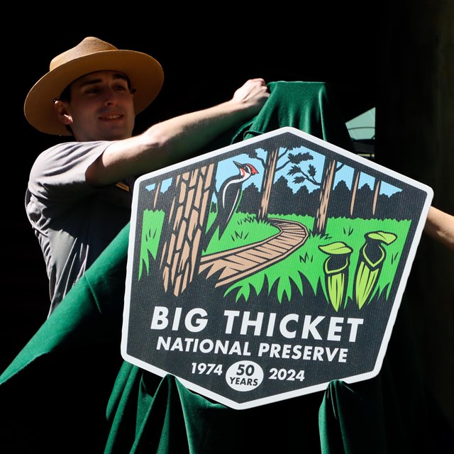 2 park rangers unveiling a Big Thicket's 50th anniversary logo on an easel outdoors.