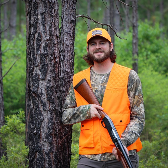 A smiling man wearing orange vest and hat, holding a shotgun, while leaning against a pine tree.
