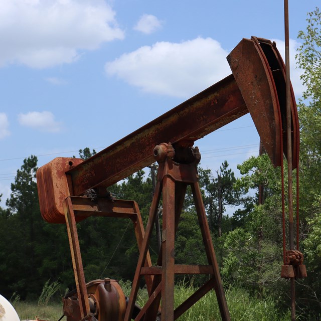 A rust-colored oil pumpjack on the edge of a forest under a blue sky with a flew clouds.