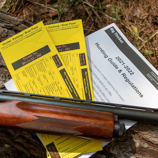 A shotgun laying on top of 2 yellow hunting permits and a copy of the hunting regulations.