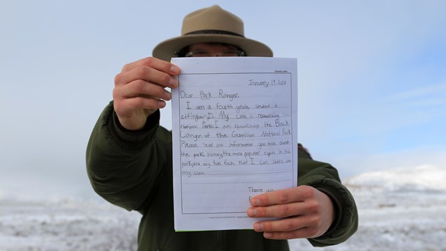 A park ranger wearing a beige hat and green jacket holds out a handwritten letter 