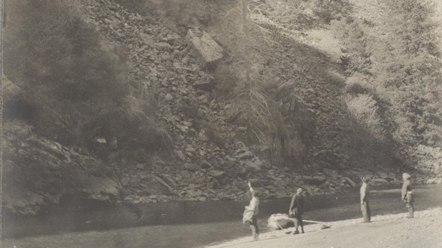 Historic image of four men standing on a riverbank looking up at steep canyon walls.