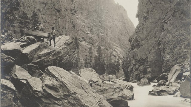 Historic image of people hauling a wooden boat from a river over large boulders.