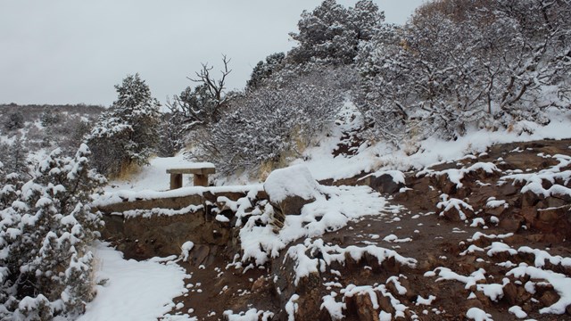 Snow covers a hiking trail, bench, and vegetation along a canyon edge