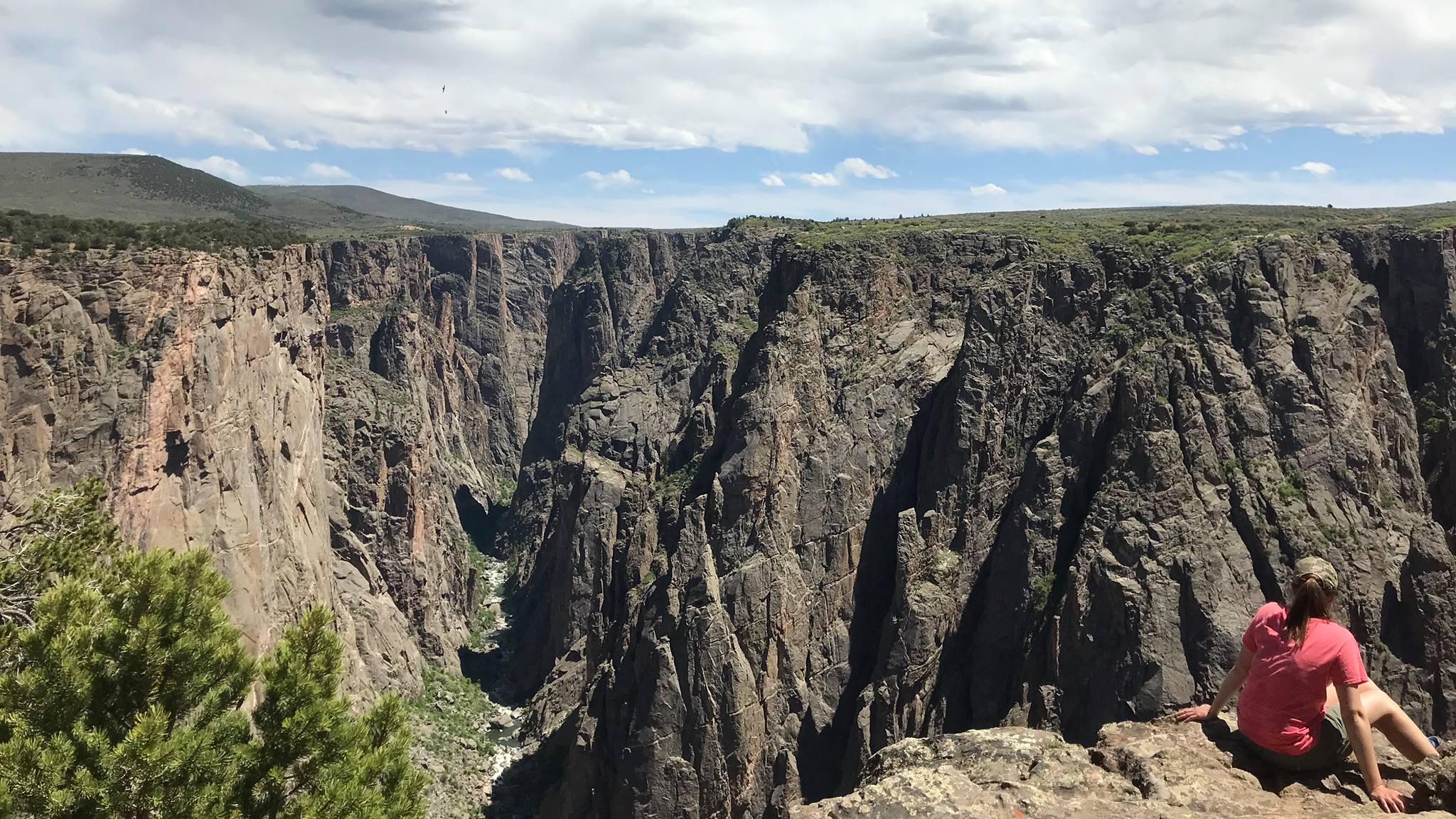 A hiker sits on a large rock overlooking a dark, steep canyon.