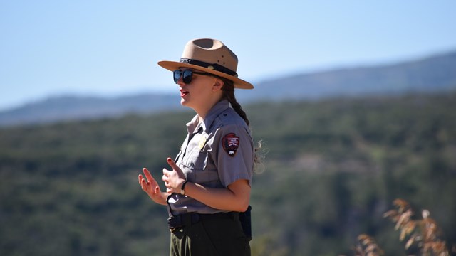 A park ranger in a uniform and flat hat stands in front of a blurry landscape.