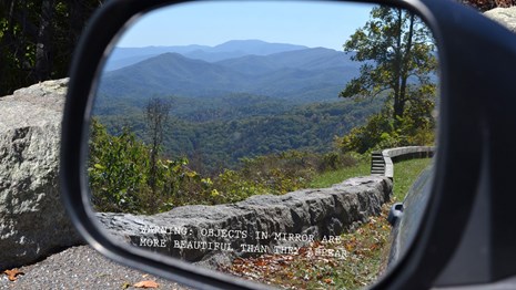 A side mirror on a car door shows ranges of mountains under a clear sky.