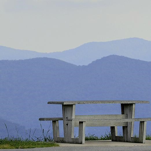 A picnic table on the edge of a grassy field, overlooking mountains.
