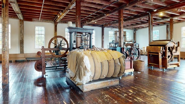 Interior of Slater Mill: Wooden floors, beams, spinning machinery, cotton bales, and sunlit windows.