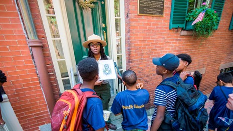 An African American Ranger standing outside of a brick house speaking to a group of students.