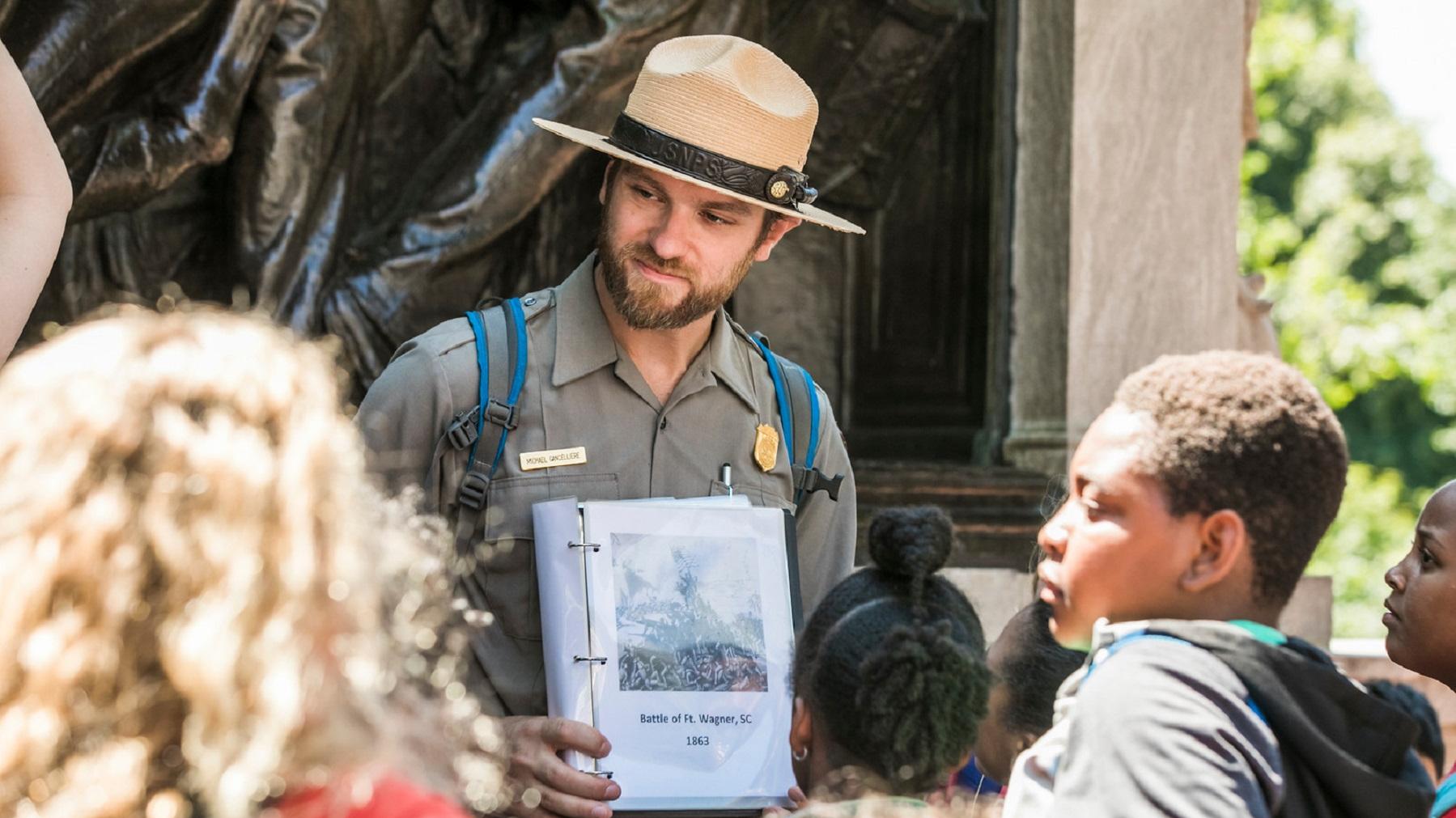An African American Ranger standing outside of a brick house speaking to a group of students.
