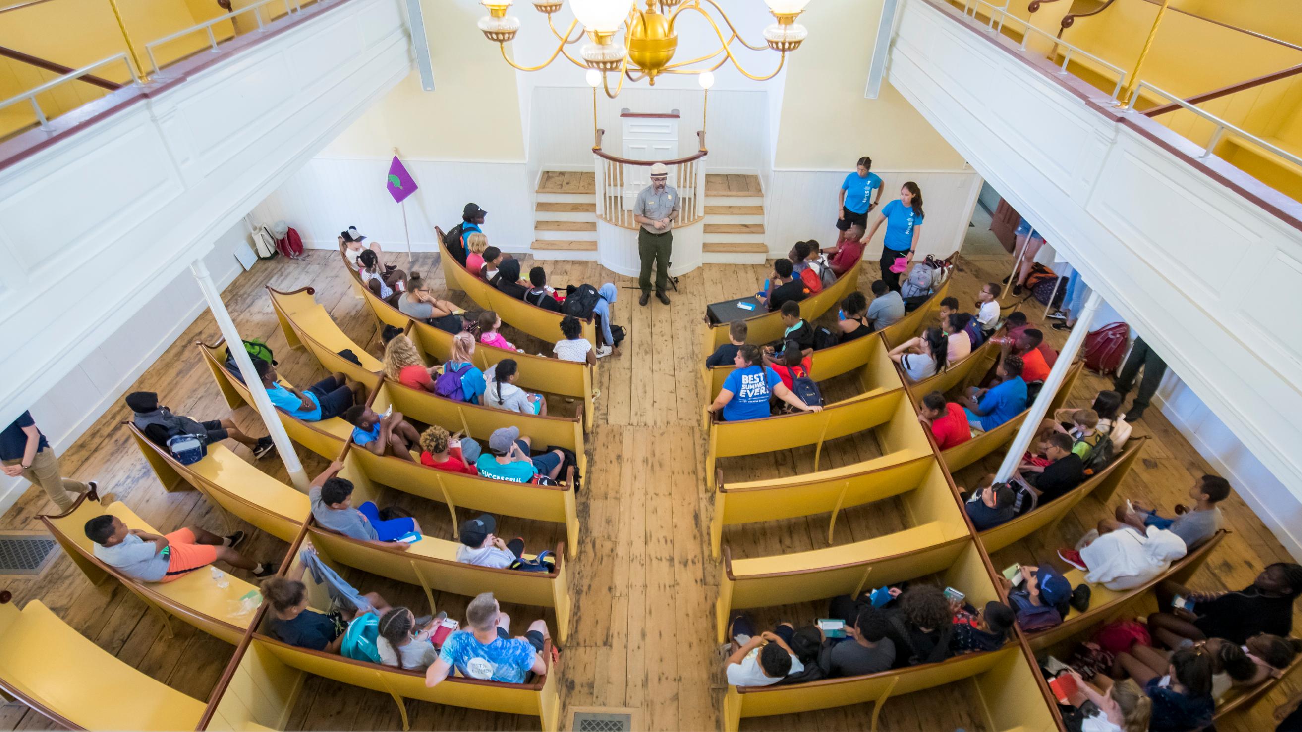 View from second story onto first floor of meeting house that has pews filled with kids.