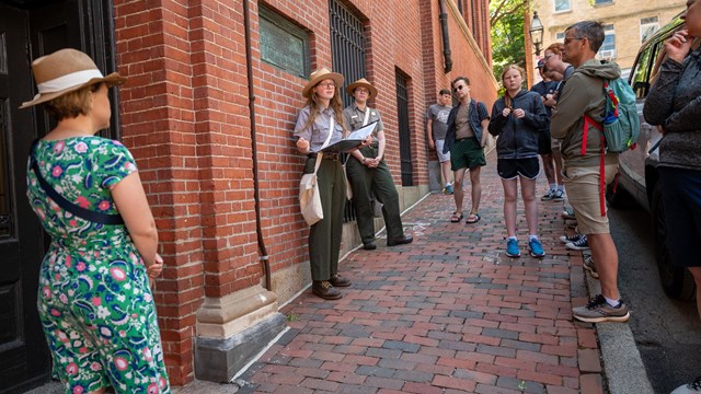 Two park rangers stand talking to a crowd of people in front of a brick building