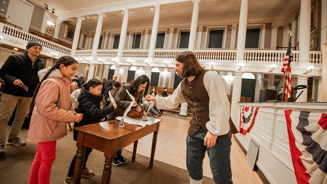 A ranger in 1800s cloths smiles at a kid signing the resolutions 