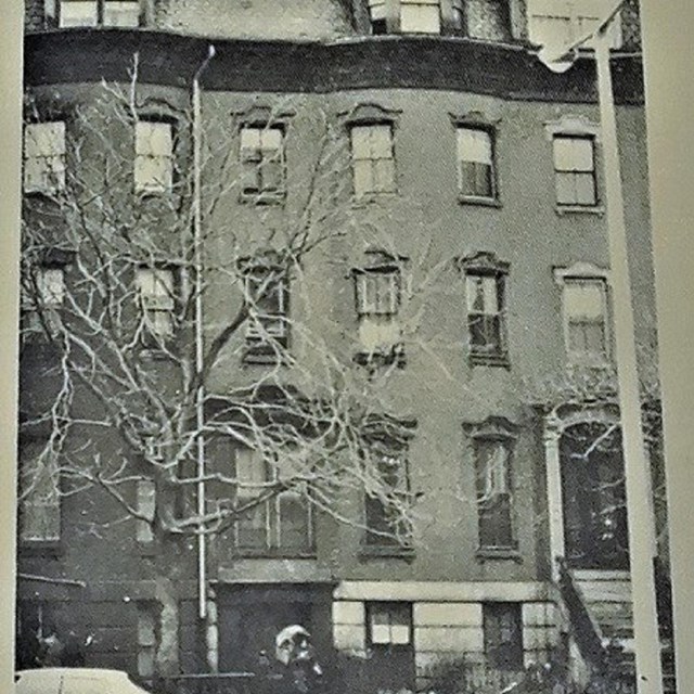 Black and white image of a five-story brick building with six windows across each floor. 