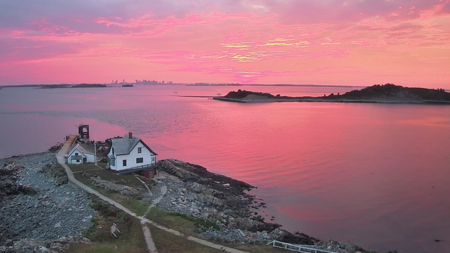 A rocky and grassy island with a few buildings overlooking other islands and the skyline at sunset