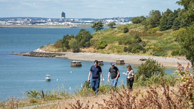 A ranger walks with two visitors along a gravel path on an island overlooking the harbor