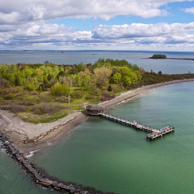 aerial view of an island with trees and some bushes. A pier and a rocky beach in foreground. 