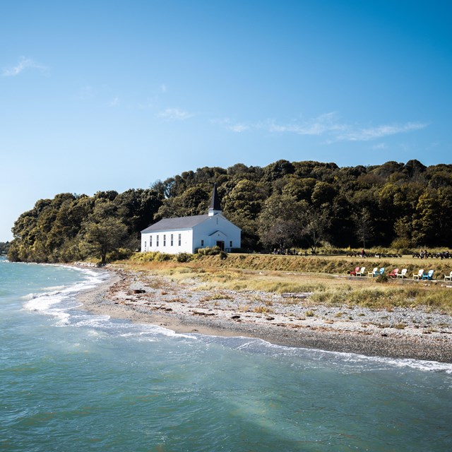 An island coastline with a pebble beach and low grass. A white chapel and trees in the background.