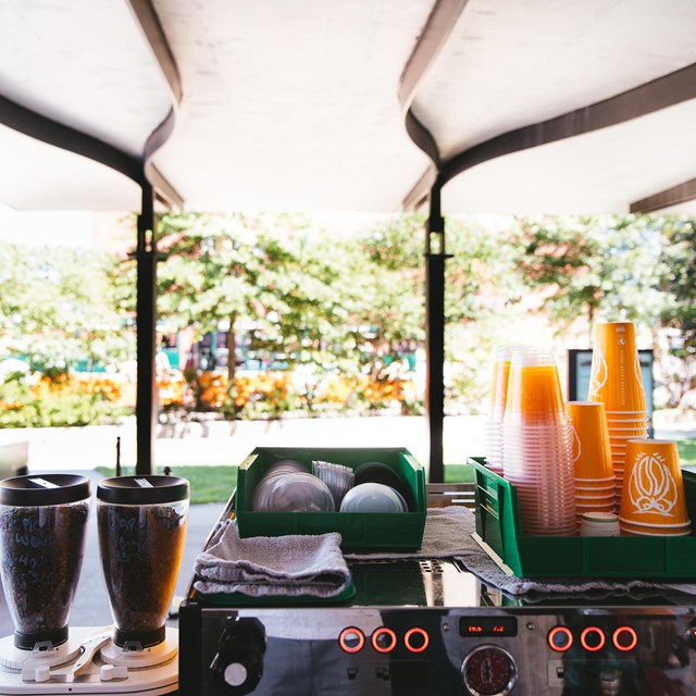 Coffee cups and coffee equipment in foreground, trees blurred in background.