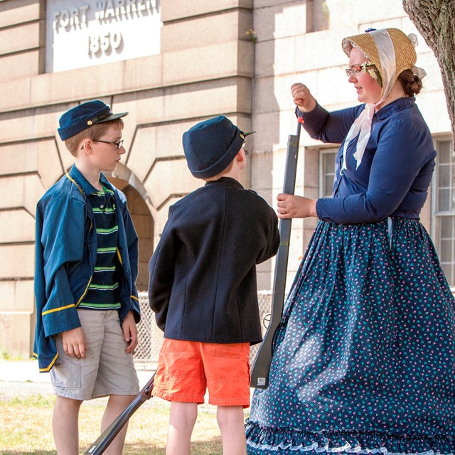 A woman wearing a Civil War period dress instructs three young boys.