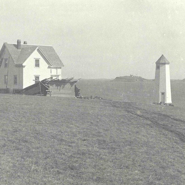 A white octagonal lighthouse next to two small white houses on an open grass area.