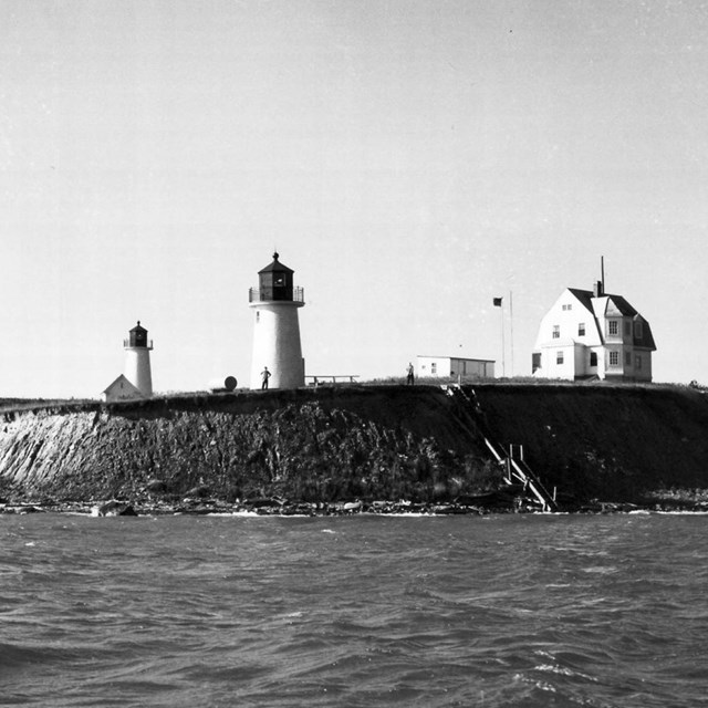 View from the ocean of two twin white lighthouses near a large white house on an island. 