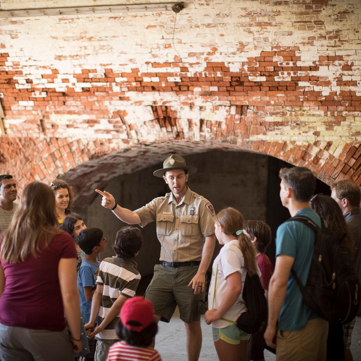 A uniformed ranger speaks to a crowd of mixed ages in a brick hallway. 