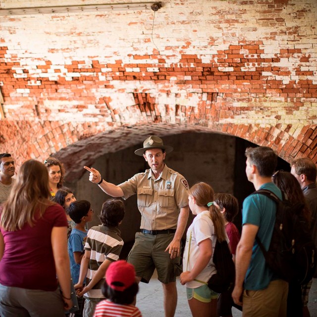A uniformed ranger speaks to a crowd of mixed ages in a brick hallway. 