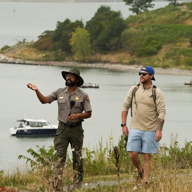 A uniformed ranger walks with a guest on a grassy path on an island, boats in the water